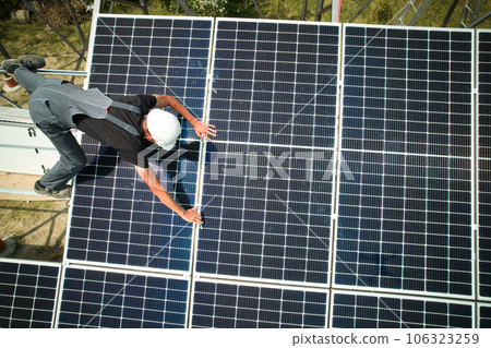 Aerial view of man technician installing solar panels to high steel platform at sunny day. Engineer wearing uniform and helmet. Concept of alternative energy. 106323259