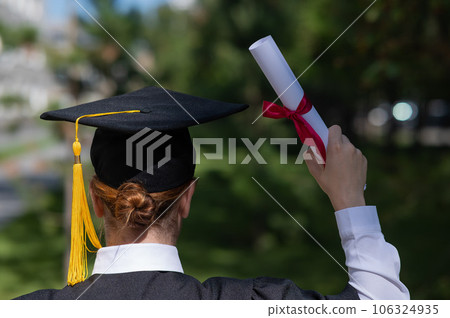 Rear view of a caucasian woman in a graduate gown holding a bundle with a diploma and standing near the university.  106324935