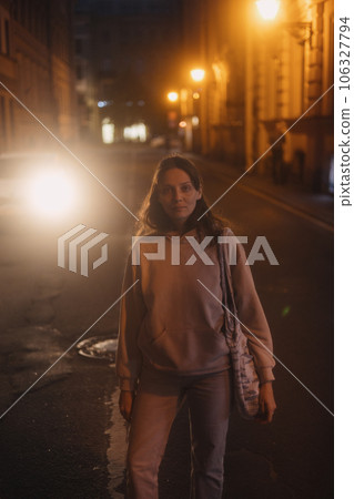 young thoughtful woman stands on road in beige hoodie. walk in evening in light of lanterns, 106327794