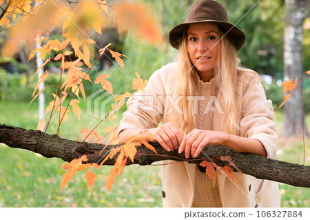 Beautiful young stylish blonde woman white coat and dark hat stands in autumn forest and look camera Beautiful young stylish blonde woman white coat and dark hat stands in autumn forest and look camera 106327884