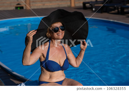 An elderly woman in a huge straw hat and sunglasses is sunbathing by the pool. Retiree on vacation. 106328355