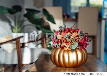 Autumn bouquet of bright artificial flowers in a golden pumpkin vase on the wooden coffee table with modern living room interior background. Hygge home fall decor for a cozy home atmosphere. 106328470