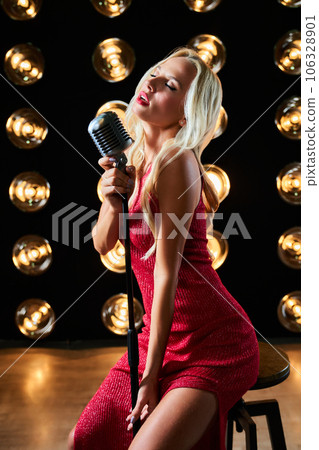 Inspired female singer singing song with closed eyes on stage. Side view of involved blonde woman performing romantic song, while sitting on barstool with vintage microphone. Concept of music. 106328901