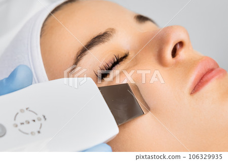 Specialist in rubber gloves removes rashes on woman face with ultrasonic device. Appearance care and spa service in light premise concept 106329935