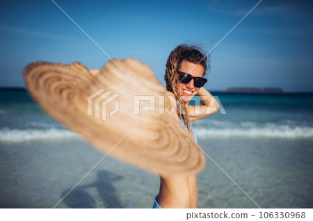 Portrait of a beautiful smiling woman with straw hat in hand standing on the beach. 106330968