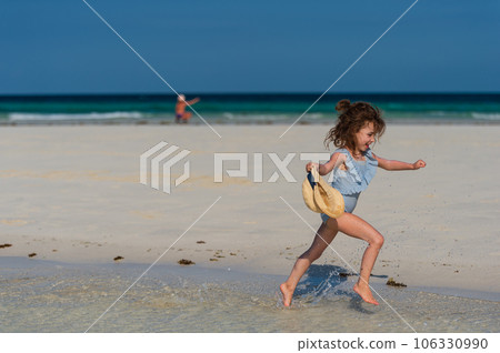 Side view of a beautiful little girl running on the beach in a swimsuit and a straw hat in hand. Side view of a beautiful little girl running on the beach in a swimsuit and a straw hat in hand. 106330990
