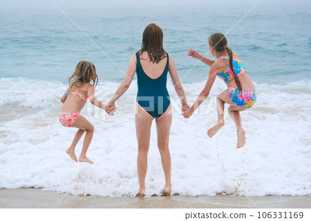 Mother and children playing on the ocean beach. Family enjoying the ocean. Mother holds girls's hands and they all look at the ocean together. Mother and children playing on the ocean beach. Family enjoying the ocean. Mother holds girls's hands and they all look at the ocean together. 106331169