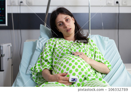Pregnant patient wearing medical oximeter, looking at camera while waiting to receive medical examination from doctor. Woman expecting child and preparing to give birth in hospital maternity room 106332789