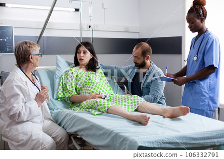 Diverse medical team discussing with pregnant woman before caesarean surgery in hospital ward. Patient with pregnancy lying in bed having contractions, being comforting by husband in maternity clinic 106332791