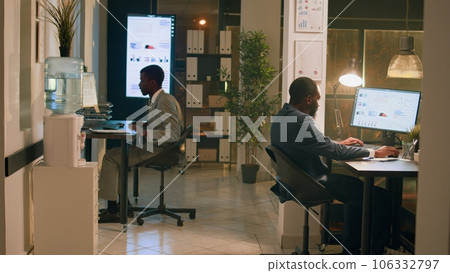 African american employees having fun at work during nightshift, holding race with desk chairs. Relaxed coworking businessmen enjoying overnight stay in office, playing together 106332797
