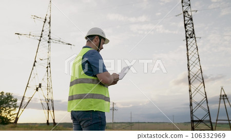 Electrician conducts diagnostic of power transmission lines with tablet in field. Engineer with tablet works at electrical power plant in field. Skilled engineer controls work of electric power lines 106332969