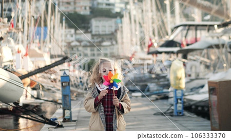 Little girl blows on a toy rainbow windmill by the sailboats in marina. Clean wind energy and peace concepts Little girl blows on a toy rainbow windmill by the sailboats in marina. Clean wind energy and peace concepts 106333370