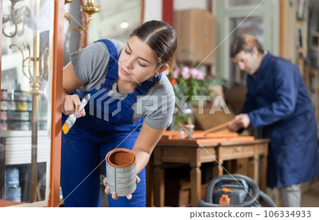 Young female furniture workshop worker repainting vintage glass cabinet 106334933