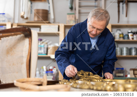Skilled old furniture repair specialist in uniform restoring Victorian mirror frame during workday in workshop Skilled old furniture repair specialist in uniform restoring Victorian mirror frame during workday in workshop 106335018