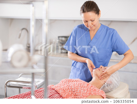 Elderly woman lies in chair during appointment with cosmetologist and enjoys facial massage Elderly woman lies in chair during appointment with cosmetologist and enjoys facial massage 106335054