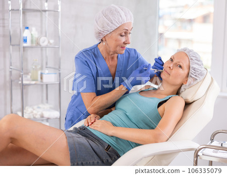 Mature woman doctor gives injection and performs procedure of lip augmentation of female patient 106335079