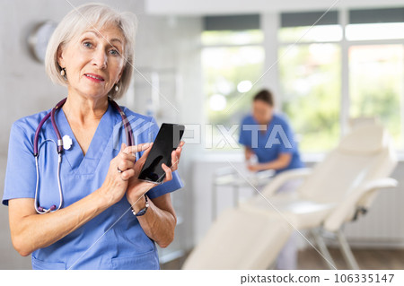 Mature woman doctor holds phone in hands and points finger at dark screen of cellphone 106335147