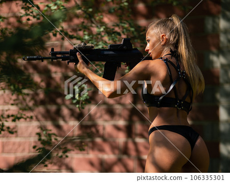 A caucasian woman in a bikini holds a machine gun against a brick wall. 106335301