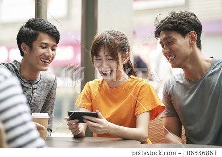 three young asian adults sitting in outdoor coffee shop using cellphone together three young asian adults sitting in outdoor coffee shop using cellphone together 106336861