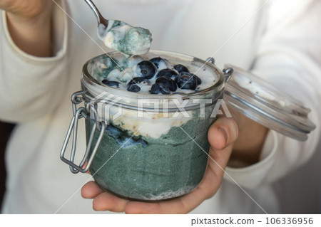 Unrecognizable woman eating Glass jar with natural organic homemade yogurt and blueberries spirulina chlorella superfood super powder blue algae sea weed. Immunity-boosting ingredients. Concept of 106336956