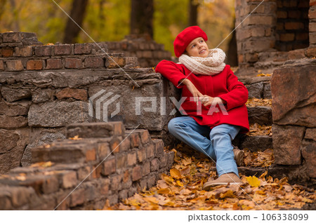 Smiling caucasian girl in a red coat and beret sits on a brick wall on a walk in autumn. Smiling caucasian girl in a red coat and beret sits on a brick wall on a walk in autumn. 106338099