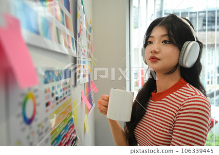 Young creative woman in headphone drinking coffee and looking at with color swatch samples chart on wall 106339445