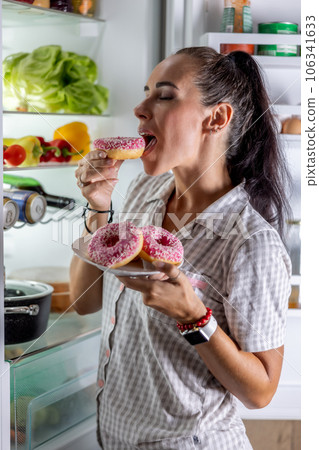 Hungry brunette in pajamas enjoys sweet donuts late at night by the open refrigerator Hungry brunette in pajamas enjoys sweet donuts late at night by the open refrigerator 106341633