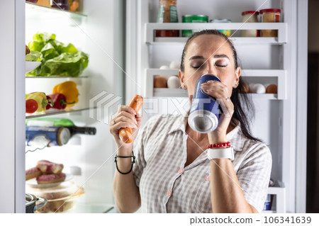 Hungry woman in pajamas drinks beer and eats sausages by the refrigerator at night 106341639