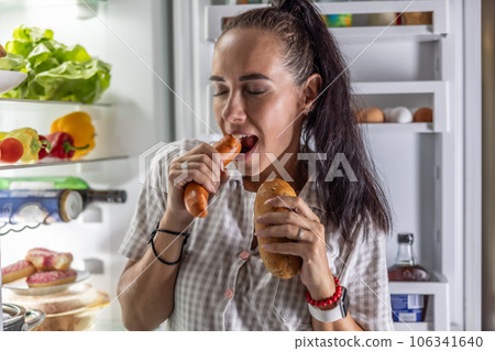 Hungry woman in pajamas eats sausages with bread by the refrigerator at night 106341640