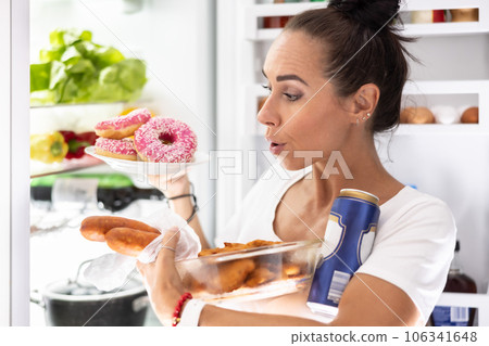 On Friday evening, a very hungry woman in pajamas chooses snacks, snitzels, donuts and a can of beer from the refrigerator 106341648