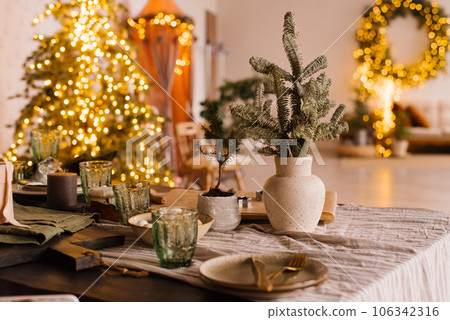 Stylish decoration for Christmas. Fir branches in a ceramic vase against the background of lights on a set table Stylish decoration for Christmas. Fir branches in a ceramic vase against the background of lights on a set table 106342316