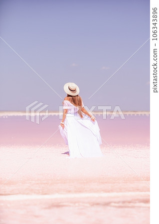 Woman in pink salt lake. She in a white dress and hat enjoys the scenic view of a pink salt lake as she walks along the white, salty shore, creating a lasting memory. 106343896