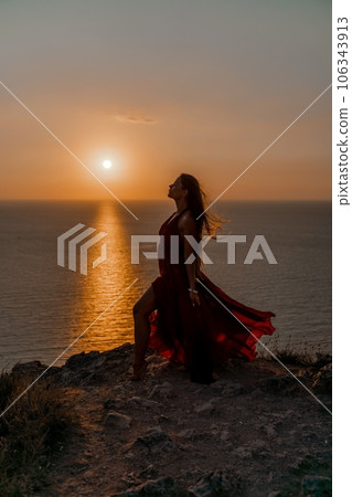 Woman sunset sea red dress, back view a happy beautiful sensual woman in a red long dress posing on a rock high above the sea on sunset. 106343913