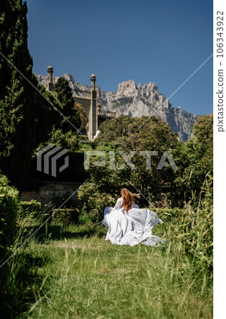 a beautiful woman with long brown hair and long white dress stay along a path along beautiful bushes in the park rear view 106343922