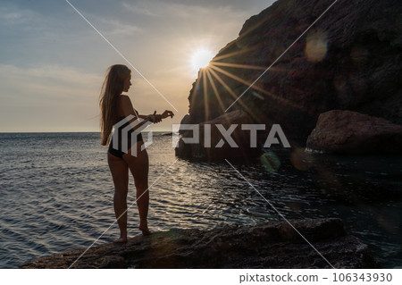 Woman tourist enjoying the sunset over the sea mountain landscape. Sits outdoors on a rock above the sea. She is wearing jeans and a blue hoodie. Healthy lifestyle, harmony and meditation Woman tourist enjoying the sunset over the sea mountain landscape. Sits outdoors on a rock above the sea. She is wearing jeans and a blue hoodie. Healthy lifestyle, harmony and meditation 106343930