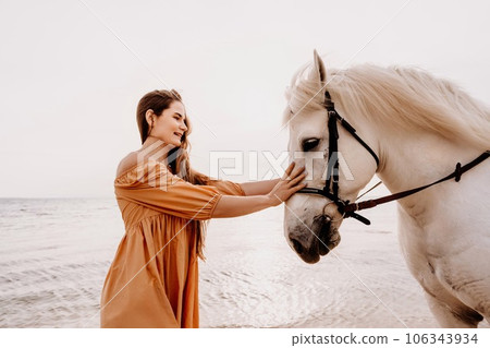 A white horse and a woman in a dress stand on a beach, with the sky and sea creating a picturesque backdrop for the scene. A white horse and a woman in a dress stand on a beach, with the sky and sea creating a picturesque backdrop for the scene. 106343934