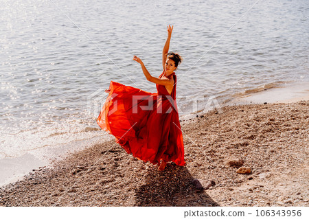 Woman red dress sea. Female dancer in a long red dress posing on a beach with rocks on sunny day. Girl on the nature on blue sky background. Woman red dress sea. Female dancer in a long red dress posing on a beach with rocks on sunny day. Girl on the nature on blue sky background. 106343956