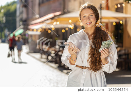 Happy teenager girl counting money dollar cash, use smartphone calculator app in urban city street 106344254