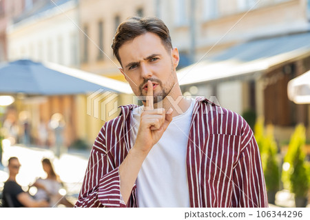 Adult man presses finger to lips makes silence hush sign do not tells gossip secret quiet in city 106344296