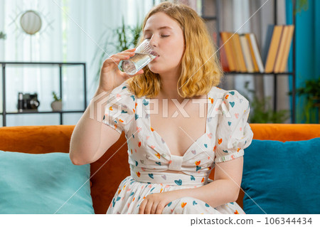 Thirsty young woman sitting at home holding glass of natural aqua make sips drinking still water 106344434