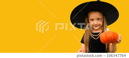 Banner with portrait of a little girl in a witch costume with a spider web pattern on his face and a pumpkin in his hands on the orange background. Copy space. 106347764