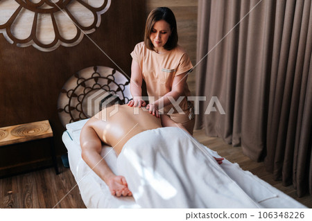 High-angle view of unrecognizable man getting back massage in spa salon lying on massage table from professional masseuse. Male client lying on stomach having full body treatment in wellness center. 106348226