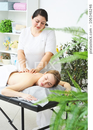 the masseur massages the back, lower back, shoulders and neck of a young woman against the background of a bright office and greenery. 106348246