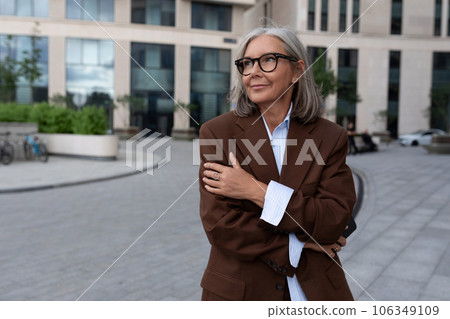 charming successful senior woman with gray hair and glasses is dressed in a brown designer suit 106349109