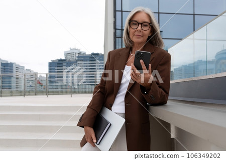 a slender senior business woman with gray hair and glasses dressed in a jacket is waiting for a 106349202
