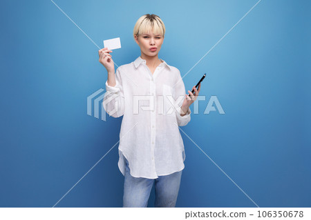 young pretty blond career woman in a white shirt holds a credit card and a smartphone. e-business 106350678