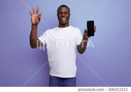 charming young african man in white t-shirt holding smartphone with mockup on studio background with 106351531
