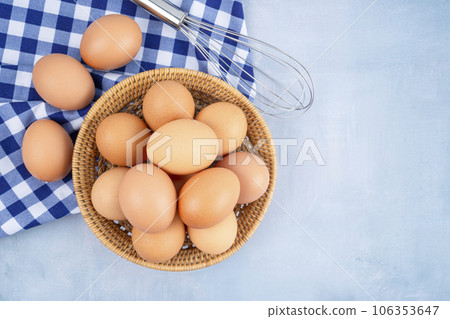 The Overhead view of brown chicken eggs in weave basket and a whisk on blue wooden background. The Overhead view of brown chicken eggs in weave basket and a whisk on blue wooden background. 106353647