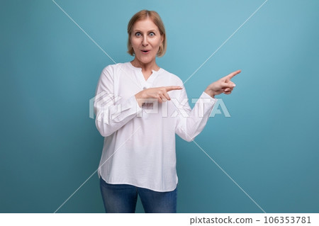 surprised blonde middle aged woman in white blouse pointing aside with finger on studio background 106353781
