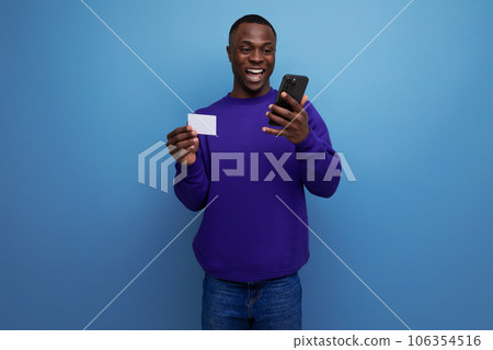 handsome young american man in blue sweatshirt pays the bill online with card and smart phone 106354516